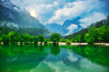 Mountain lake landscape, with lush green pine forrest,  misty clouds and sun peeking from behind a peak