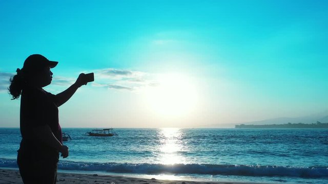 A woman alone on a shoreline of tropical beach taking photos of the beautiful afternoon sun setting over the blue water