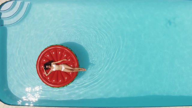 Aerial view of a woman in white bikini relaxing on big red floating mattress in the swimming pool on hot sunny day. Female enjoying summer holidays.