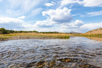 A highland river valley with yellow grass under white clouds and blue sky, The beautiful landscape of Mongolia near the city of near Karakorum. Cristal clear water in a mountain creek.
