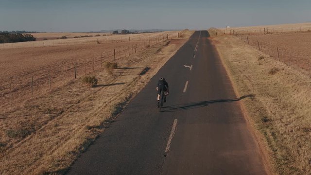 Rear view of a professional athlete cycling on long countryside highway. Aerial view shot of a sportsman cycling uphill on the rural asphalt road.