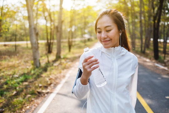 Closeup image of an asian female runner holding and drinking water from bottle after jogging in city park