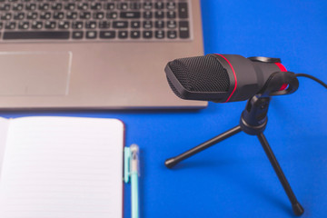 A microphone for recording sound and podcasts next to a laptop and notebook on a blue background. Desktop of blogger.