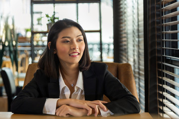 Close up charming young Asian woman in black suit sitting ans smiling see outside the window at modern office.