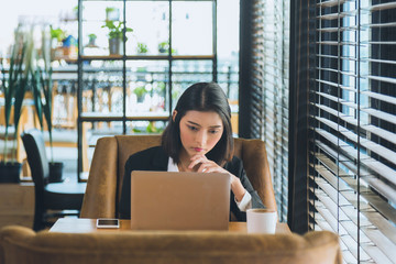 Close up charming young Asian woman in black suit sitting and touching her chin working with laptop on wooden table with cellphone and coffee cup