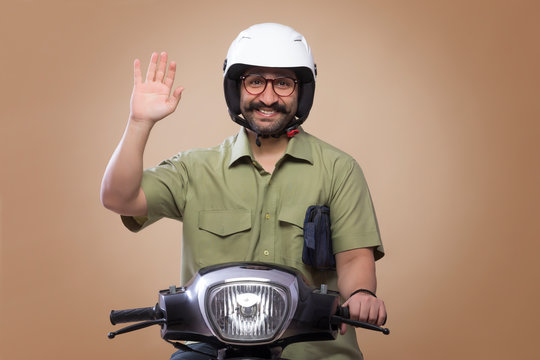 Smiling Man With A Small Bag In His Arms Riding Scooter Wearing A Helmet And Showing Palm.