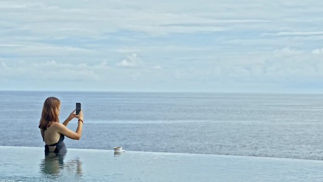 young woman in infinity pool with coffee and holding mobile phone to take picture video. pretty female relax on sea ocean horizon. take photo panorama on vacation in bali hotel exotic resort