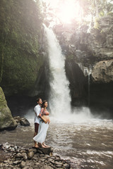 Young pregnant couple in love with amazing view of Tegenungan cascade waterfall. Morning sunlight, Happy together, pregnancy travel lifestyle. Honeymoon in Bali.