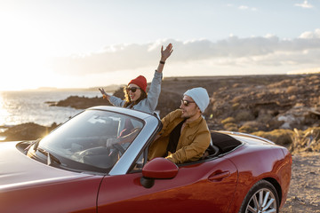 Joyful couple enjoying vacations, driving together convertible car on the rocky ocean coast on a sunset. Happy vacation, love and travel concept