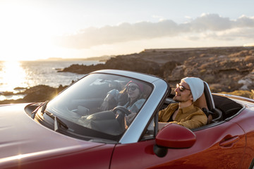 Fototapeta na wymiar Joyful couple enjoying vacations, driving together convertible car on the rocky ocean coast on a sunset. Happy vacation, love and travel concept