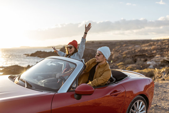 Joyful couple enjoying vacations, driving together convertible car on the rocky ocean coast on a sunset. Happy vacation, love and travel concept