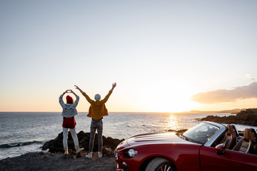Couple enjoying beautiful views on the ocean, standing together with raised hands near the car on the rocky coast, wide view from the side with copy space on the sky