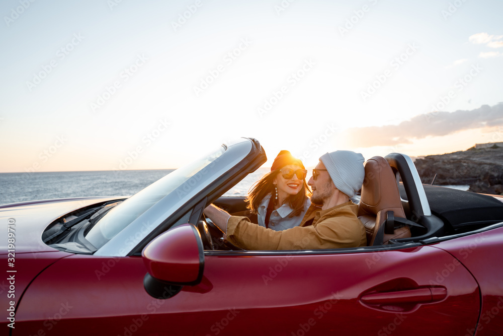 Wall mural Joyful couple enjoying vacations, driving together convertible car near the ocean on a sunset. Happy vacation, love and travel concept