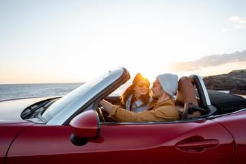 Joyful couple enjoying vacations, driving together convertible car near the ocean on a sunset. Happy vacation, love and travel concept - obrazy, fototapety, plakaty