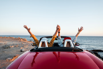 Couple driving convertible car, traveling near the ocean on a sunset, view from the backside. Happy vacations and traveling by car concept - Powered by Adobe