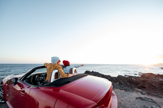 Couple enjoying beautiful view on the ocean, traveling by convertible car near the ocean on a sunset, view from the backside. Happy vacations and traveling by car concept - Powered by Adobe