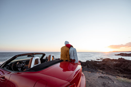 Couple enjoying beautiful views on the ocean, hugging together near the car on the rocky coast, wide view from the side with copy space on the sky