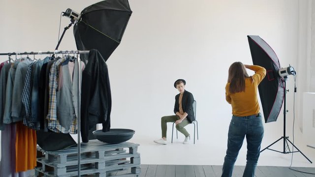 Slow motion of female photographer taking pictures of male model in studio using professional camera, equipment and clothing on hangers is visible.