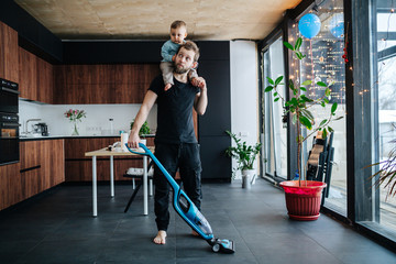 Young father vacuums apartment floor with his baby riding on his neck