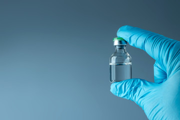 Hand in a blue glove holds a bottle with a vaccine close-up on a gray background, medical concept vaccination subcutaneous injection, dose. Disease treatment immunization. - obrazy, fototapety, plakaty