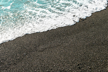 Shallow volcanic black sandy beach and pebbles, blue water waves and white foam washes the coastline with wet dark sand on the sea coast. Ocean shore natural background, copy space.