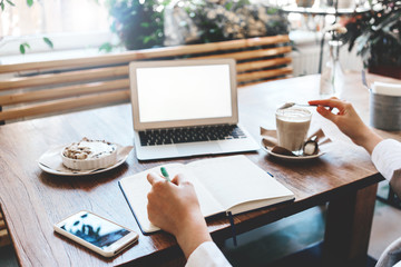 Close-up of woman's hands making notes in her note book. Workplace in city coffee house. Laptop, mobile phone, cake and cup of coffee on the table