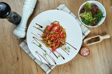 Appetizing wok noodles with tomatoes and chicken in a white plate on a wooden background. Restaurant style street-food. Close up view