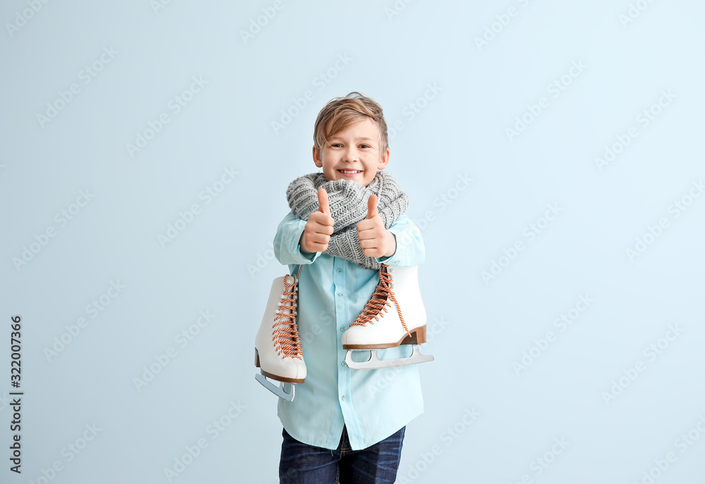 Wall mural Cute little boy with ice skates showing thumb-up against color background