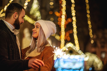 Christmas, winter holidays conception: young happy hugging couple with sparkler, models looking at each other, posing in street of European city. Outdoor night portrait. Copy, empty space for text