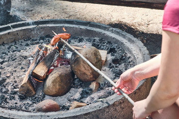 Obraz premium Woman fries sausages at the campfire outdoor. Woman Having Barbecue On Outdoor.