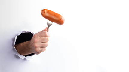 Man's hand holding hot boiled wiener on fork through ripped hole in white paper wall. Street food....