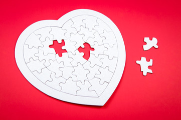 White jigsaw puzzle in the shape of a heart missing two pieces sitting nearby on red background