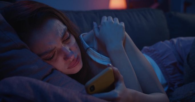Close up of lonely young woman lying on sofa and using smartphone at night. Depressed millennial female starring at mobile phone screen and thinking. Concept of painful brokeup.