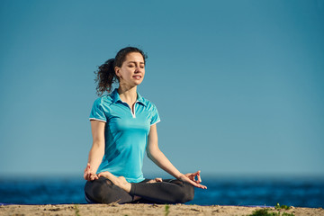 Relaxed young woman meditating while sitting on sandy shore in lotus pose
