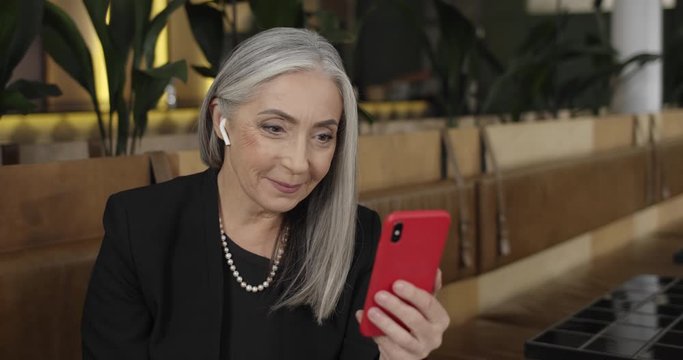 Older good looking woman in earpods waving to smartphone camera whilehaving video conversation. Mature elegant businesswoman sitting in cafe and having video conference.