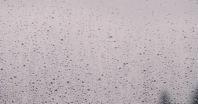 High-contrast raindrops slowly sliding down a window with grey cloudy out of focus background.
