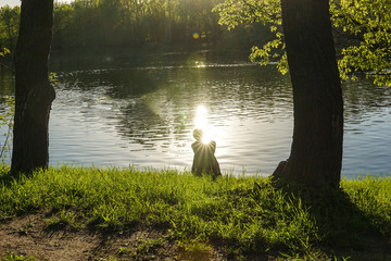 Silhouette of a person with sun glare on his shoulder, sitting on the shore of the pond. - 322020166