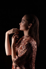 Young pretty woman in a sparkling dress with sequins stands with face in profile. Lady posing with her hands and elegant fingers on a black background. Fashion portrait of a girl