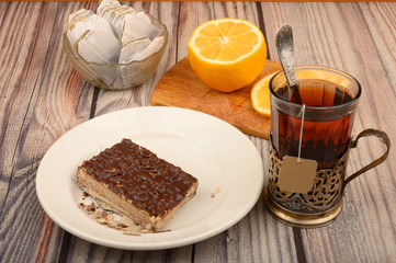 Tea in a faceted glass in a vintage Cup holder, tea bags in a glass vase, sliced lemon on a Board and a piece of waffle cake on a white plate on a wooden background. Close up.