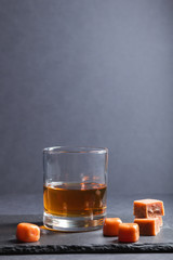 Glass of whiskey with caramel candies on a black stone slate board on black background. Side view, copy space.