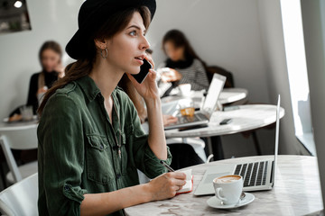 Woman in hat talking phone in office and planning her week, concentrated busy woman. Freelancer, entrepreneur, work in cowering space, public place, modern people lifestyle, business online concept.