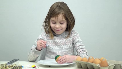 The model of European appearance is a girl of 6-7 years old. The girl is focused on coloring chicken and quail eggs for the holiday. In front of the child, a dozen eggs in a box.