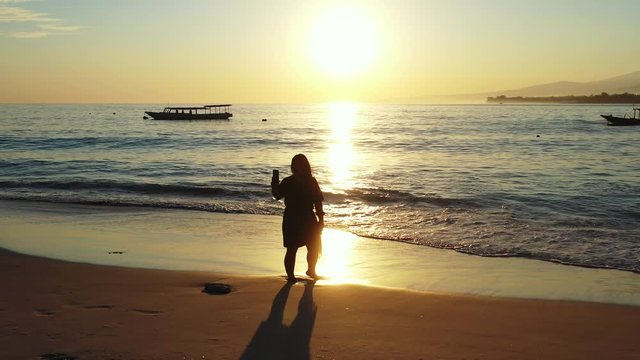 Girl taking selfie photos with smartphone on empty beach at sunset golden hour with orange sky reflecting on sea surface