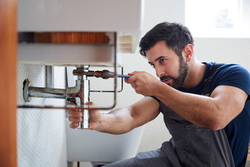 Male Plumber Using Wrench To Fix Leaking Sink In Home Bathroom