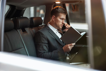 young businessman having phone conversation holds his planner while sitting in his car on the way to meetting with partners, multitasking concept