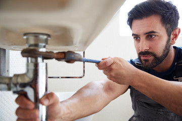 Male Plumber Using Wrench To Fix Leaking Sink In Home Bathroom - obrazy, fototapety, plakaty