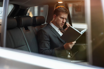 young businessman having phone conversation holds his planner while sitting in his car on the way...