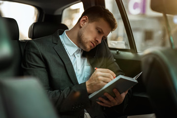 successful businessman takes notes to planner while having important phone conversation riding on...