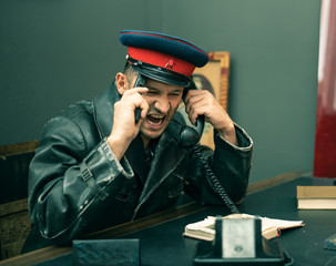 The Soviet man security officer wears cap with red star and hammer and sickle at table in office