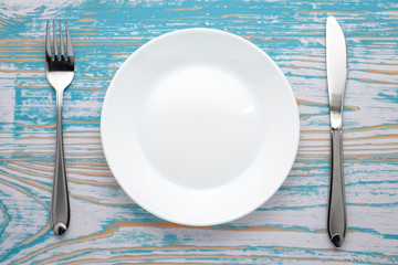 Empty white plate with silver fork and knife on blue wooden table. Dinner place setting. Top view.